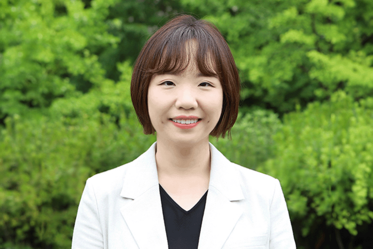 Korean woman with short, dark brown hair with bangs. She is smiling at the camera, wearing a short-sleeve ivory jacket and a black inner top. Behind her, the area is filled with bushes and shrubs.