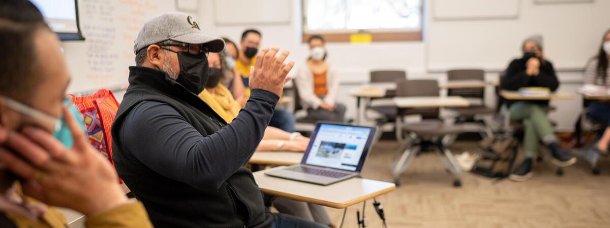 student in baseball cap and mask gestures while speaking; other students in background
