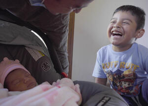 Image of man setting down a car seat carrier with a newborn sleeping in it while smiling at a laughing young child