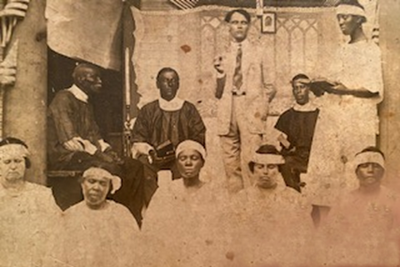Sepia photo (approximately 1920s) showing a preacher and deacons on a platform with a young woman standing to their right with an open book in her hand. Church members / choir members are in the foreground.
