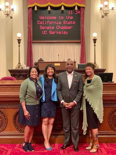 Dean Burton visiting the California State Senate Chamber, a sign behind her reads "Welcome to the California State Senate Chamber"
